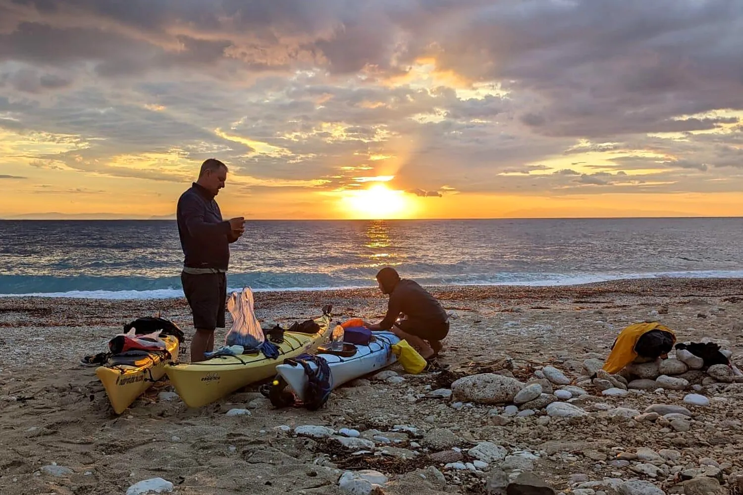 Two loaded sea kayaks on a stony Greek beach at sunrise, with two paddlers packing kit before setting out